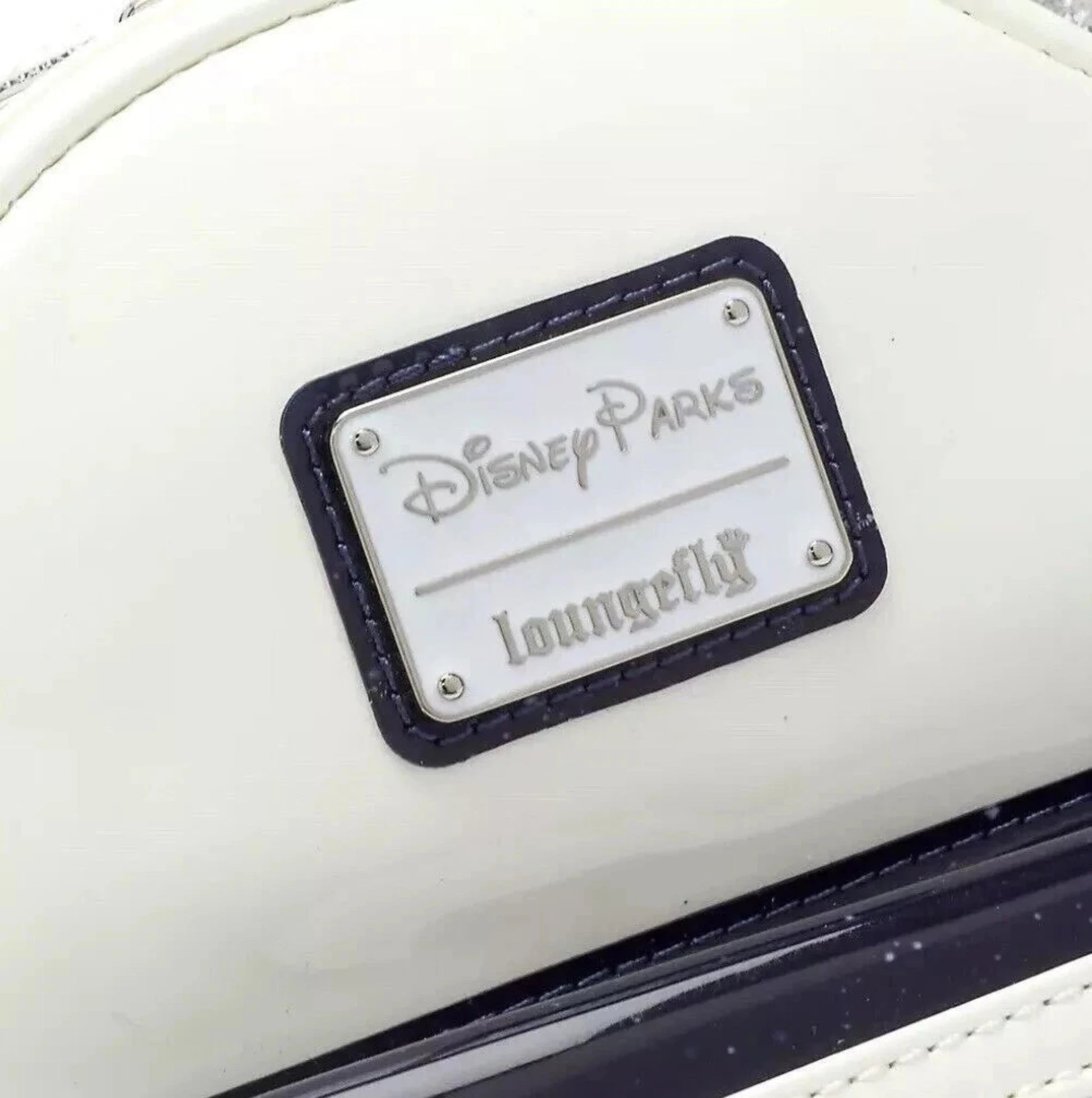 Disney Parks Loungefly Rucksack – Detailaufnahme des silbernen Logoschilds mit geprägtem „Disney Parks“ und „Loungefly“-Schriftzug auf weißem Kunstleder.